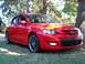 2007 Mazda 3 - This red 2007 Mazda 3 hatchback is parked on a grassy area with trees in the background.
