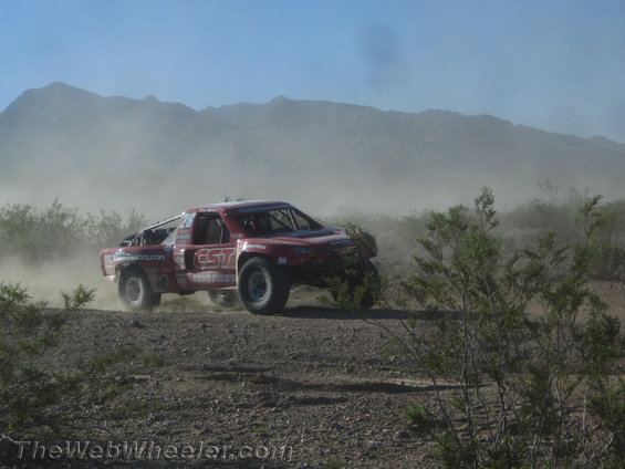 Red off-road racing truck kicking up dust on a dirt track with mountains in the background
