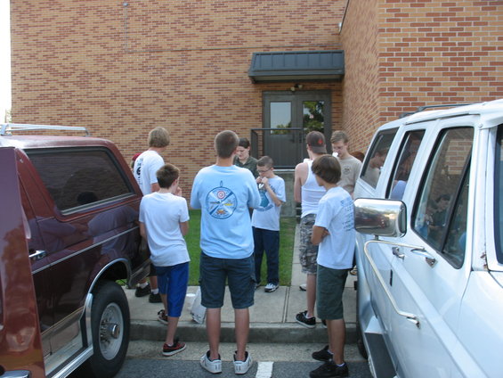Side view of a maroon SUV and a white van with several young males standing around them