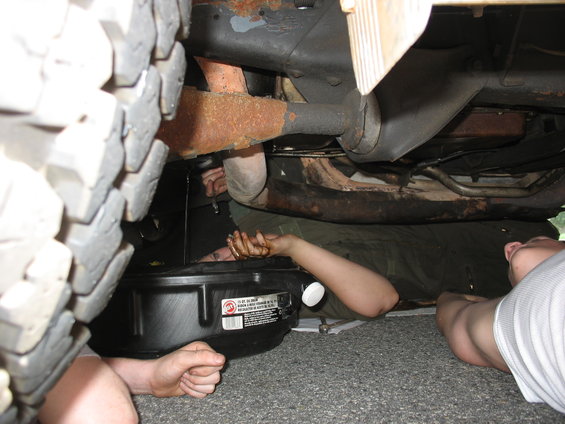 Two people are lying on the ground, working on the underside of a vehicle. One person is draining oil into a pan