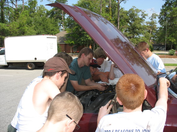 Boys gathered around a maroon car engine with the hood open