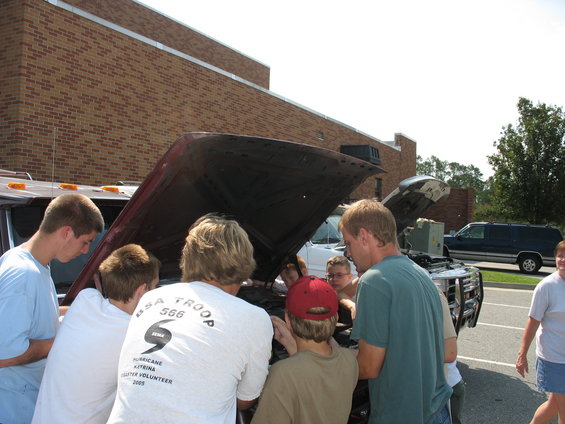 Several guys looking at the engine of a dark red pickup truck. Another van is parked behind it