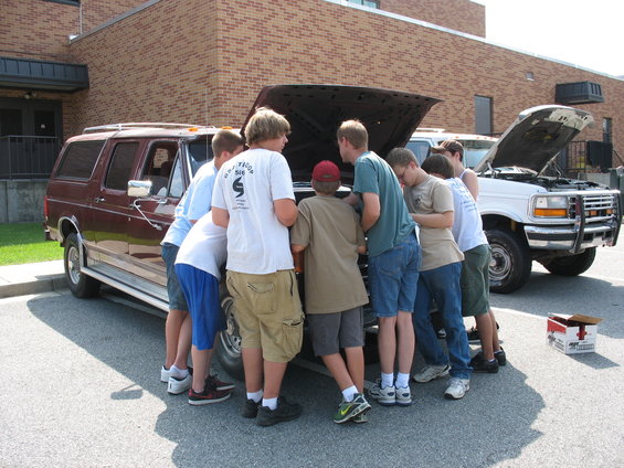 Maroon SUV and white truck with hoods open, boys gathered around engines