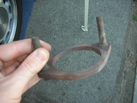 Rusty exhaust flange with two bolts held by a hand, with a car wheel and blue carpet visible