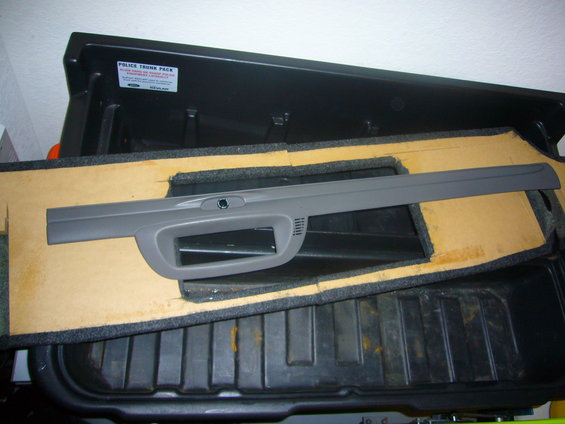 Gray plastic interior trim piece with a cutout and a small circular button, resting on foam and carpeted material inside a black plastic trunk organizer