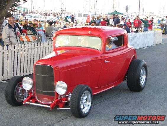 Red hot rod coupe with large rear tires, chrome wheels, and exposed front suspension, viewed from a 3/4 front angle