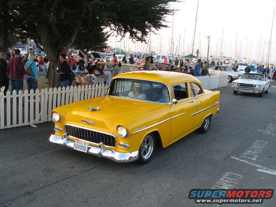 Yellow 1955 Chevrolet Bel Air driving past a white picket fence with a marina in the background. A white convertible is visible behind it