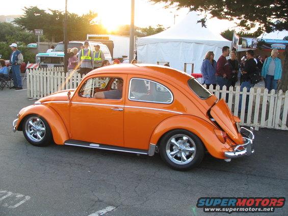 Orange Volkswagen Beetle with chrome wheels, rear engine lid open, parked near a white fence and tents