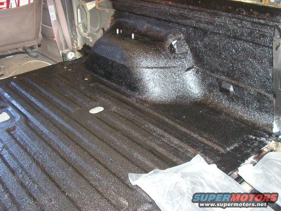 Interior of a vehicle bed with black spray-on liner applied. Seats are visible in the background