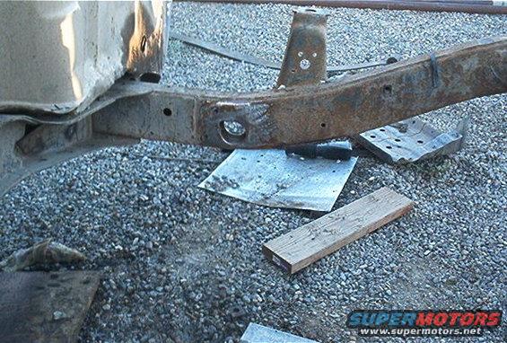 rusted vehicle frame section with holes and brackets, resting on gravel