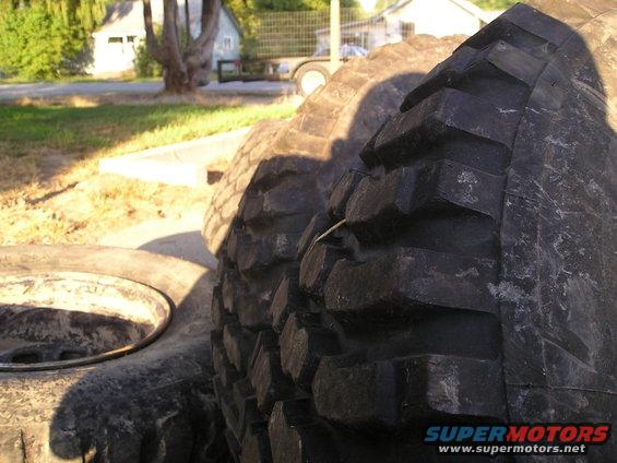 Stack of large off-road tires, one with visible tread detail. Supermotors.net logo in corner