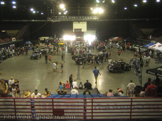 Indoor car show with many vehicles and people milling around. Spectators sit in bleachers in the foreground