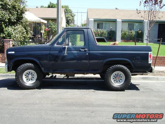 Dark blue Ford Bronco parked on street, side view. Houses and trees in background