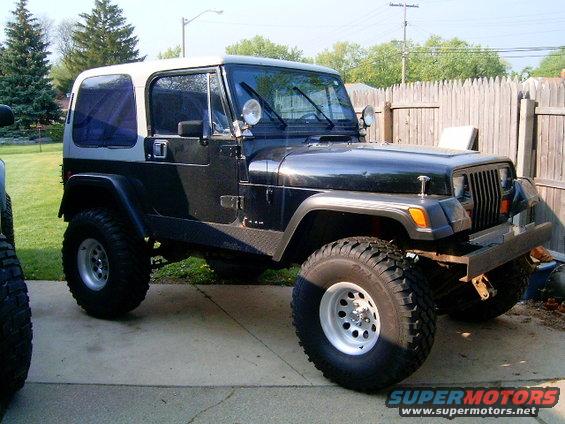 Black Jeep Wrangler with a white hardtop, parked in a driveway next to another vehicle. It has large off-road tires and a custom bumper