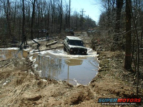 Dark SUV navigating a muddy water obstacle; 1970s era vehicle