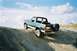 1990 Ford Bronco - This 1990 Ford Bronco is captured in a three-quarter front view as it navigates an incline on a dirt path under a cloudy sky.