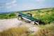 1990 Ford Bronco - 1990 Ford Bronco navigating an off-road incline with a scenic backdrop of water and vegetation.