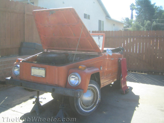 Orange Volkswagen Thing on jack stands, hood open, showing spare tire. License plate reads 'VINTAGE AUTO'