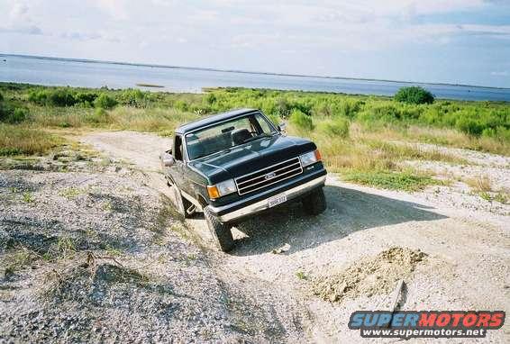 1990 Ford Bronco driving on a dirt road near a body of water