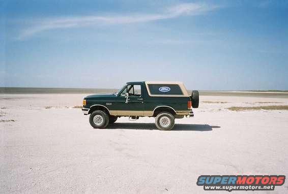 Green 1990 Ford Bronco with tan trim and a spare tire on the back, parked on a sandy, flat landscape under a blue sky