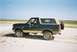 1990 Ford Bronco - A 1990 Ford Bronco with a tan top and spare tire mounted on the rear is seen on a sandy terrain under a clear blue sky.