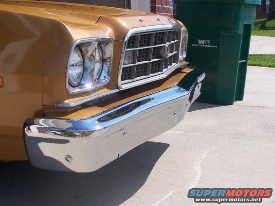 Gold car's front end with chrome bumper and grille. Green trash bin to the right. Supermotors.net logo at bottom right