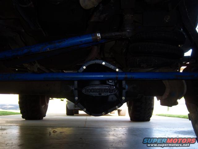 Underneath a vehicle showing a differential cover labeled 'ROCKCRUSHER' and part of another vehicle in the background