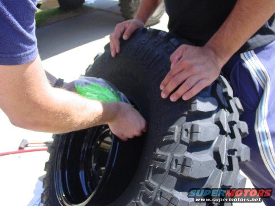 Two people are working on a large off-road tire and black rim