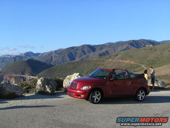 Red convertible PT Cruiser parked on a scenic overlook with mountains in the background. Two people stand near the car