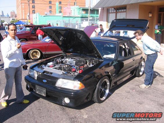 Black Ford Mustang with hood and trunk open, showing engine. Man in white shirt stands next to it. Red car behind