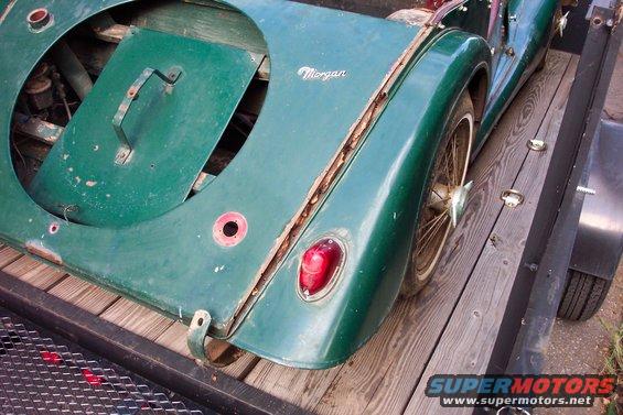 Green Morgan car's rear section on a wooden trailer, showing a spare tire cover, taillight, and wire wheel