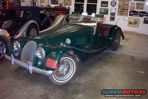 1964 Morgan 4/4 convertible sports car in dark green with red interior, parked next to a blue convertible