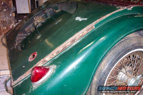 Green car trunk and rear fender with visible rust and a wire wheel. Text on bottom right reads 'SUPERMOTORS.NET'