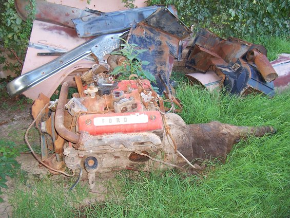 Rusty Ford V8 engine and transmission in grass, with chrome bumper and other car parts
