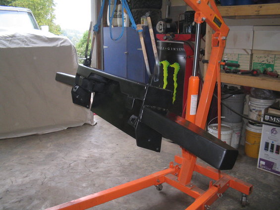 Black aftermarket bumper mounted on an orange engine hoist in a garage
