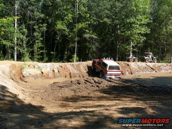 1988 Ford Bronco climbing out of a mud pit, two ATVs on the bank behind it