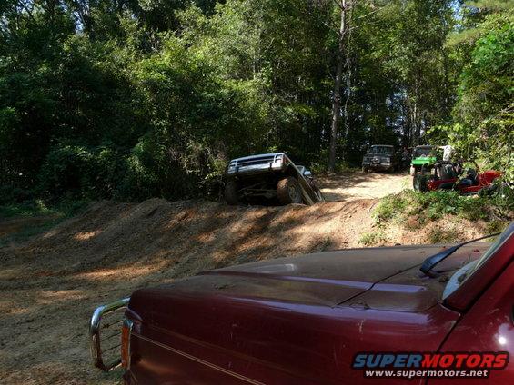 Red truck hood in foreground, white SUV on dirt ramp, green ATV and red buggy in background