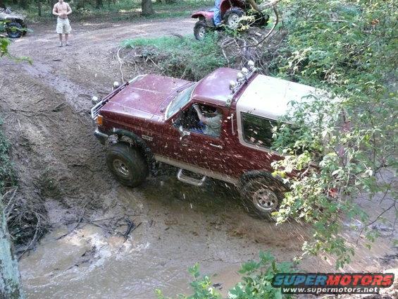 1988 Ford Bronco navigating a muddy trail with trees and an ATV in the background