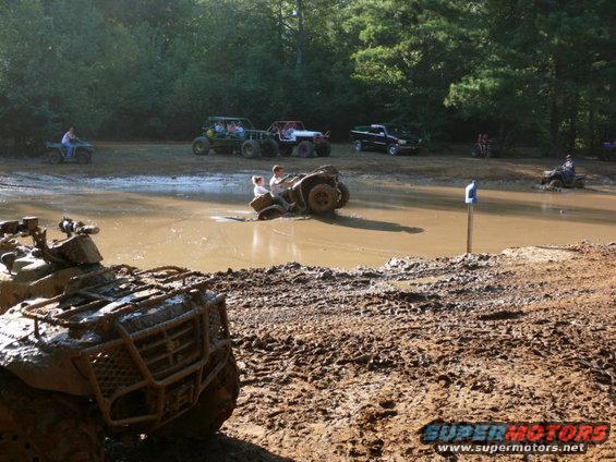 Muddy ATV in foreground, other ATVs and a truck in a waterhole with trees in background