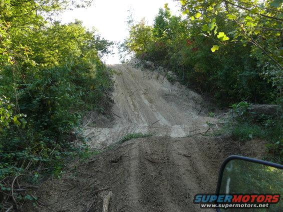Sandy off-road trail going up a steep hill with trees on either side. Tire tracks visible