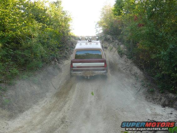 Brown and maroon Ford Bronco driving up a dirt hill, kicking up dust. Roof has four spotlights