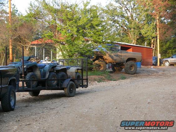 Pickup truck towing a trailer loaded with four ATVs, with a lifted mud-covered truck parked nearby
