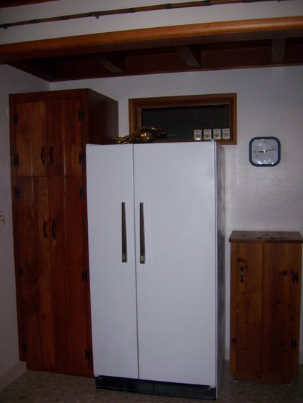 Tall wooden cabinet, white refrigerator, and small wooden cabinet against a wall with a clock and a small window