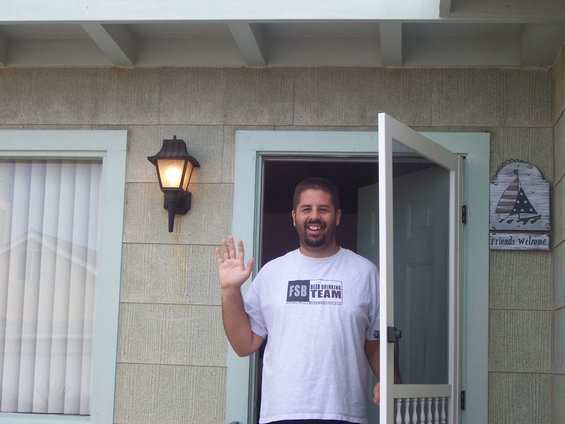 Man in a t-shirt waves from a doorway of a house with a "Friends Welcome" sign
