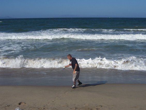 Man in dark t-shirt and shorts walks on sandy beach near ocean waves, picking up trash