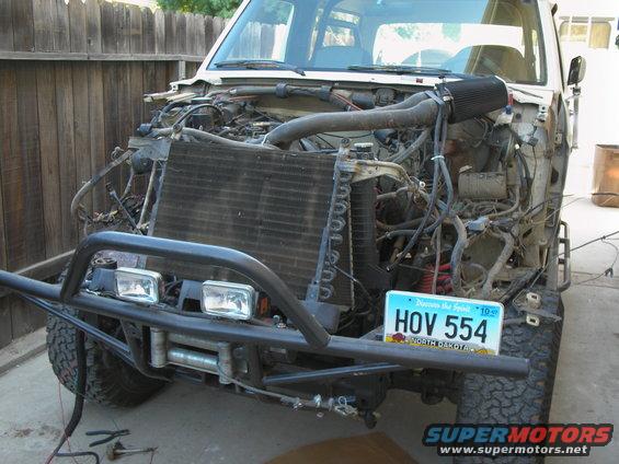 Front of a white Ford Bronco with its hood open, showing the engine and radiator. It has a custom bumper and winch