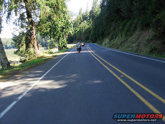 Three motorcycles ride down a two-lane road through a wooded area