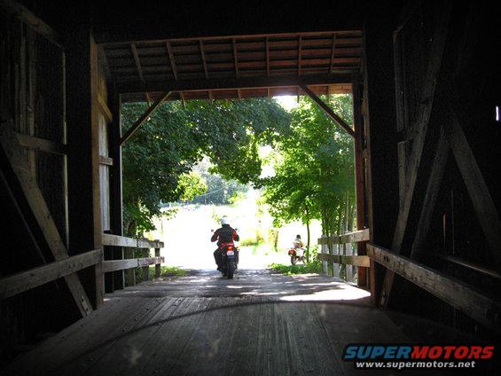Two motorcycles ride out of a wooden covered bridge into a sunny, tree-lined road