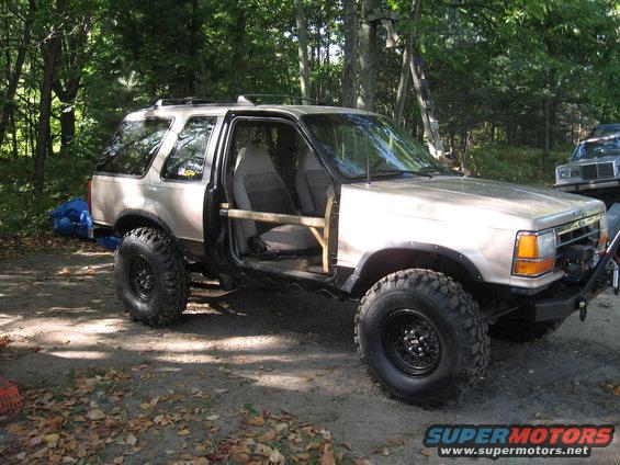 Modified SUV with large tires and no doors, parked in a wooded area. Another vehicle is visible in the background