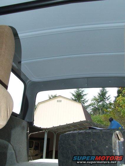 View from inside a vehicle, looking out past a spare tire and a barn. Another vehicle is visible under a carport
